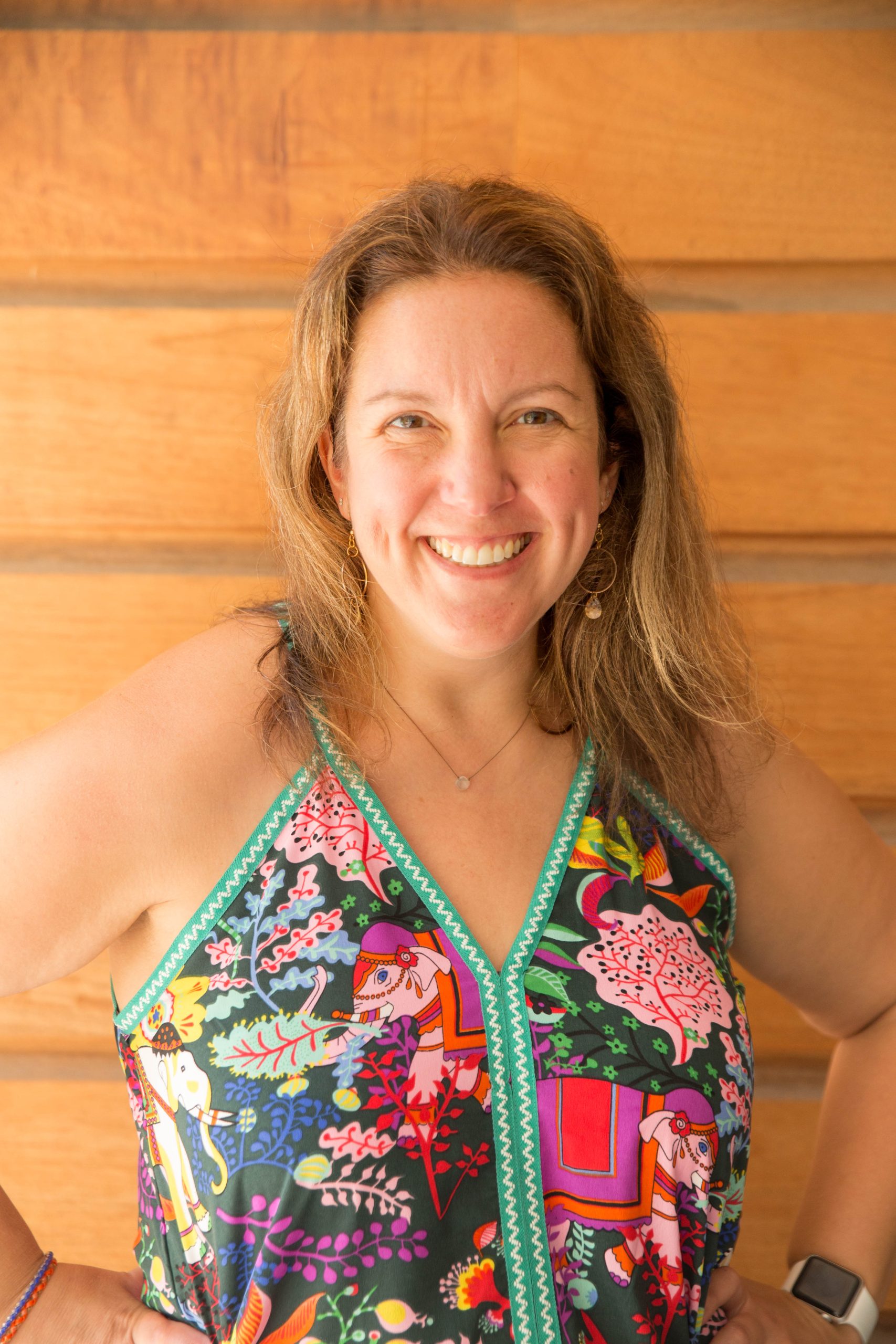 A photo of the author Megan Birch-McMichael, a white woman standing against a wooden backdrop with a multicolored dress.